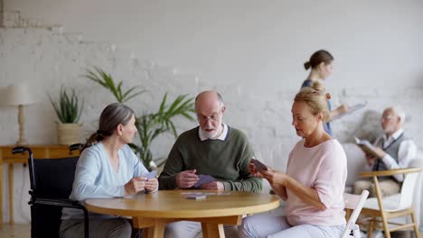 Tracking-Aufnahme-von-drei-Senioren,-die-Karten-spielen,-die-am-Tisch-im-Pflegeheim-sitzen.-Junge-Krankenschwester-im-Gespräch-mit-männlichen-alten-Patienten-Lesen-Buch-auf-Sofa-im-Hintergrund