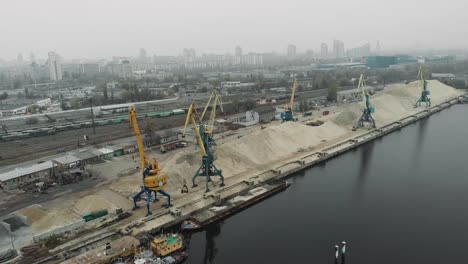 Industrial-part-of-the-city-with-cargo-construction-cranes-and-old-cargo-railway-tracks.-Extraction-of-sand-from-barge-to-pile-of-sand-with-cargo-trucks