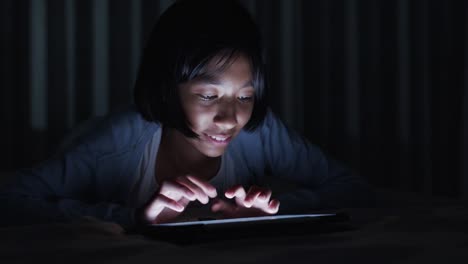 Asian-little-girl--touching-keyboard-on-modern-digital-tablet-in-the-bed-at-night