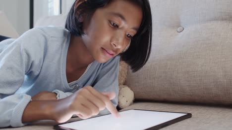 Asian-of-little-girl-touches-the-white-digital-tablet-screen-and-smile-while-lying-on-the-sofa-in-the-living-room-at-home.