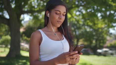 Camera-panning-over-the-young-woman-listening-to-music-on-earphones-texting-messaging-on-smartphone-in-the-park---happy-girl-on-her-phone-in-the-park-on-a-sunny-day