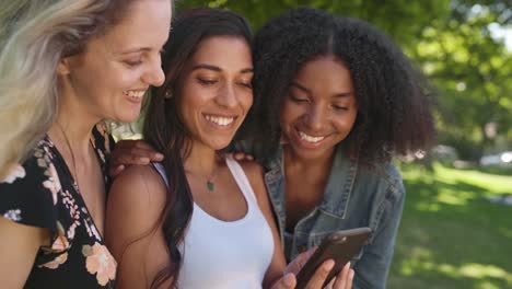 Cheerful-three-female-diverse-friends-hanging-out-in-the-park-using-mobile-phone-and-sharing-digital-content-on-social-media-smiling-and-happy---looking-at-the-phone-app