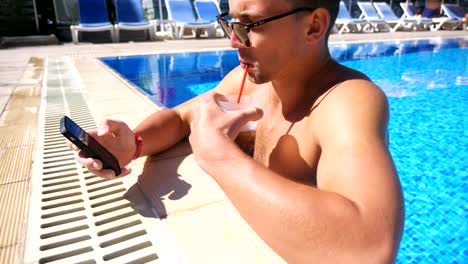 Handsome-man-using-phone-at-pool-of-hotel-and-refreshing-cold-cocktail.-Young-guy-in-sunglasses-drink-beverage-making-photo-for-social-network.-Man-relaxing-on-resort-during-summer-vacation.-Slow-motion