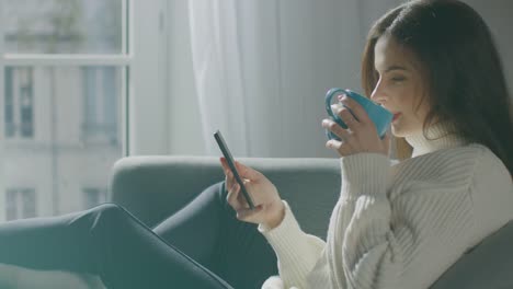 Schöne-junge-Frau-mit-Smartphone,-trinkt-Tee,-während-auf-dem-Stuhl-sitzen.-Sensual-Girl-Wearing-Sweater-Surfs-Internet,-Beiträge-auf-Social-Media,-teilt-Bilder.-Seitenansicht-Zeitlupe