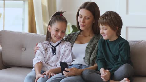 Glückliche-junge-Mama-unterrichtet-Kinder-mit-Handy-sitzen-auf-dem-Sofa