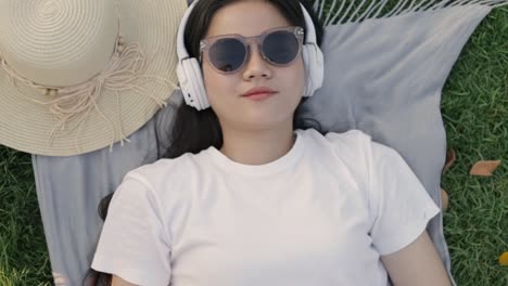 Smile-of-portrait-asian-woman-wearing-sunglasses-and-headphones-while-lying-on-the-floor-grass-outdoors-at-a-public-park-on-the-beautiful-sunset.