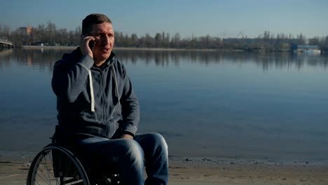 Man-in-wheel-chair-speaking-on-mobile-phone-near-river,-faith-in-future