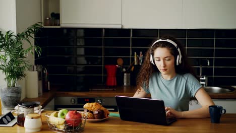 positiven-glücklich-kaukasischen-ziemlich-lockig-Teenager-Mädchen-sitzen-am-Tisch-im-Chat-mit-jemandem-auf-Laptop-Musikhören-im-Bug-Kopfhörer-in-Küche-im-Haus