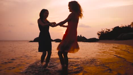 dos-chicas-baila-felices-cogidos-de-la-mano-en-la-playa-al-atardecer-en-cámara-lenta