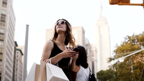 Feliz-atractivo-caucásica-joven-con-bolsas-de-la-compra-de-gafas-de-sol-mirando-hacia-arriba,-usando-la-aplicación-para-smartphone-en-Nueva-York