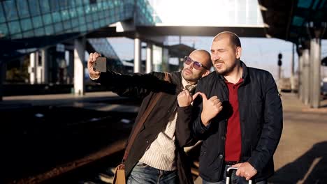 Two-men-on-the-platform-of-the-railway-station-waiting-for-the-arrival-of-the-train-and-take-pictures-selfie-on-a-smartphone.