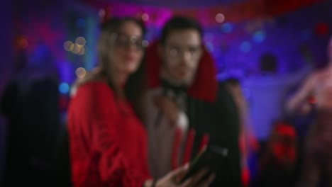 Halloween-Costume-Party:-Seductive-She-Devil-and-Handsome-Count-Dracula-Taking-Selfie-for-Social-Networks-with-a-Smartphone.-In-the-Background-Group-of-Monsters-Having-Fun,-Dancing-Under-Disco-Ball.