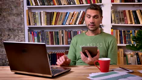 Businessman-sitting-in-front-of-the-laptop-and-having-conversation-on-a-tablet.-Bookshelves-on-the-background