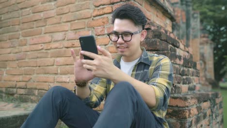 Traveler-Asian-man-using-smartphone-checking-social-media-while-relax-after-spending-holiday-trip-at-Ayutthaya,-Thailand,-Male-enjoy-his-journey-at-amazing-landmark-in-traditional-city.