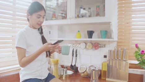 Jovencita-asiática-adolescente-bebiendo-café-caliente-usando-teléfono-inteligente-escuchando-música-y-comprobando-social-en-la-cocina-en-casa.-Estilo-de-vida-mujer-relajarse-en-la-mañana-en-el-concepto-de-casa.