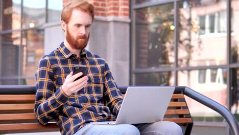 Redhead-Beard-Young-Man-Using-Smartphone-and-Laptop,-Sitting-on-Bench