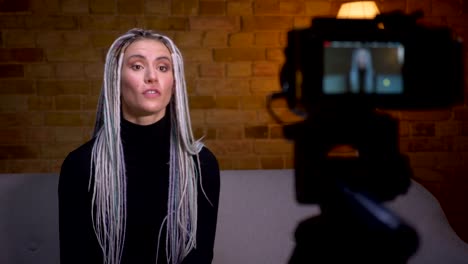 Closeup-shoot-of-young-attractive-female-fashion-blogger-with-blonde-dreadlocks-streaming-live-talking-on-camera-and-gesturing-sitting-on-the-couch-indoors