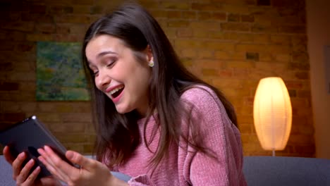 Closeup-shoot-of-young-pretty-brunette-caucasian-female-browsing-on-the-tablet-and-smiling-happily-in-a-cozy-apartment-indoors