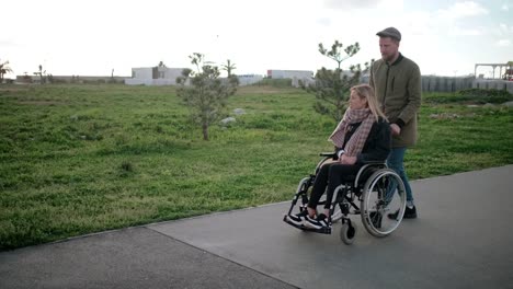 Man-is-rolling-wheelchair-with-young-blonde-woman,-walking-together-in-park