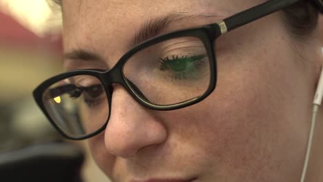 Close-up-of-eyes-of-attractive-smiling-redhead-woman-with-glasses-reflection,-freckles-and-red-hair-watching-internet-video-on-smartphone-sitting-at-bus-stop-in-street,-during-sunny-summer-in-Paris.