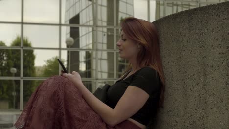 Woman-using-smartphone-Sitting-in-business-district,-texting-with-mobile-telephone-outside,-typing-text-message-on-cell-phone.-Paris,-Red-hair.-Social-media.-Smiling.-4K-UHD-Slow-Motion-Profile-view.