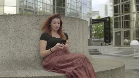 Attractive-woman-using-smartphone-Sitting-in-business-district,-laughing-while-texting-with-mobile-telephone-outside,-typing-text-message-on-cell-phone.-Paris,-Red-hair.-Social-media.-Smiling.-4K.