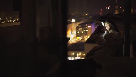 Woman-in-bathrobe-using-smartphone-at-night-time-in-hotel-suite