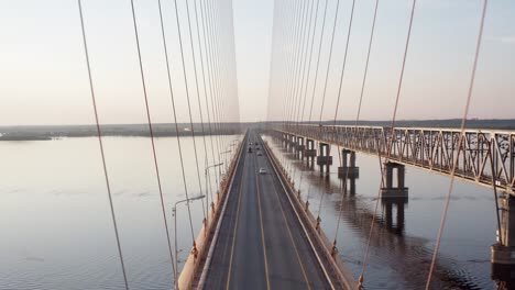 Luftfliegen-über-und-durch-eine-Kabelbrücke,-die-in-der-untergehenden-Sonne-über-dem-Fluss-steht.-4k