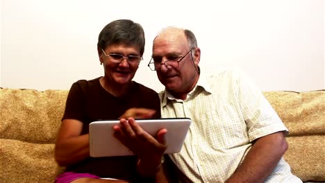 An-elderly-couple-is-sitting-on-a-sofa-at-home-and-watching-photos-on-a-tablet-pc,-smiling-and-talking-actively.