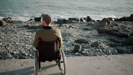 Alone-male-wheelchair-user-is-sitting-calmly-on-seacoast-and-relaxing,-back-view