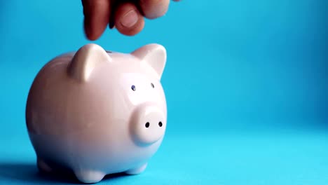 Man's-hand-putting-coins-in-piggy-bank-of-pig-shape-on-blue-background