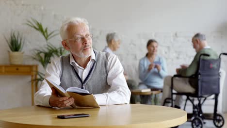 Rack-focus-of-three-elderly-people,-two-senior-women-and-disabled-old-man-in-wheelchair,-playing-cards-in-assisted-living-home.-Intelligent-old-man-in-eyeglasses-reading-book-and-thinking-at-table