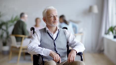 Seguimiento-de-la-toma-media-de-un-anciano-discapacitado-en-gafas-sentado-en-silla-de-ruedas-mirando-hacia-otro-lado-y-pensando-en-un-asilo-de-ancianos,-otros-pacientes-de-edad-en-segundo-plano