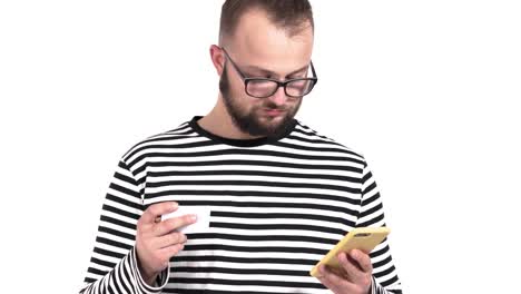 Close-up-of-a-confused-young-man-with-beard-using-his-cellphone.-He's-holding-business-card-in-his-hand-and-checking-information-in-gadget.-Social-Networks.-Apps.-Smartphones.