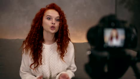 Portrait-of-red-headed-young-girl-vlogger-is-talking-in-front-of-camera-recording-video-for-online-blog-for-her-followers,-smiling-and-gesturing.-Woman-is-wearing-jeans-and-white-sweater.-Accelerated-video.-Camera-on-foreground,-blurred