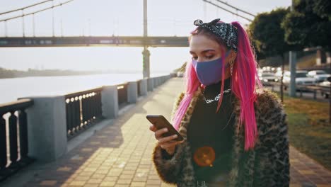 Young-hipster-lady-in-informal-outfit-and-protective-mask-is-using-smartphone-while-standing-at-embankment