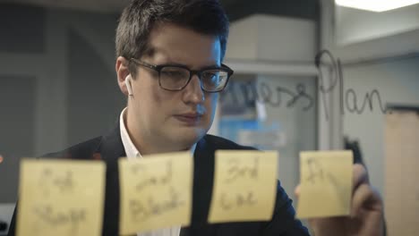 Young-boss-businessman-writing-plans-on-the-sticky-notes-on-glass-wall