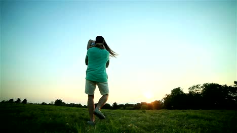 Young-man-spins-and-holds-happy-woman-in-arms-twirling-together-at-sunset-backlit-grass-POV.-Boyfriend-rotates-carrying-smiling-long-hair-girlfriend-in-sunny-evening-summer-meadow.-Love-togetherness