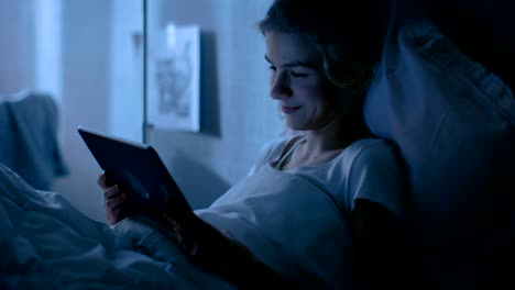 Young-Teenage-Girl-Lies-in-Her-Bed-at-Night-With-Tablet-Computer.-Watches-TV-Show-and-Smiles.