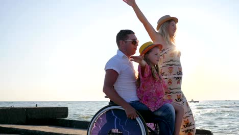 invalid-at-summer,-family-play-together-on-background-blue-sky,-little-girl-sitting-on-daddy-in-wheelchair-with-outstretched-arms