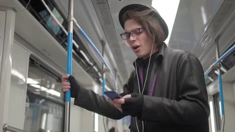 Young-stylish-male-hipster-wearing-black-hat-and-eyeglasses-with-headphones-listening-to-music,-browsing-on-smartphone-in-public-transport,-millenial-boy-enjoying-mobile-technology-in-subway