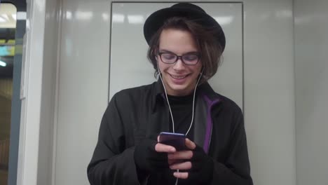 Young-stylish-male-hipster-wearing-black-hat-and-eyeglasses-with-headphones-listening-to-music,-browsing-on-smartphone-in-public-transport,-millenial-boy-enjoying-mobile-technology-in-subway