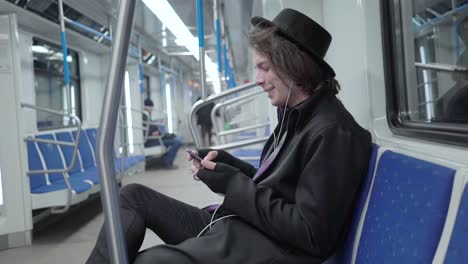 Young-stylish-male-hipster-wearing-black-hat-and-eyeglasses-with-headphones-listening-to-music,-browsing-on-smartphone-in-public-transport,-millenial-boy-enjoying-mobile-technology-in-subway