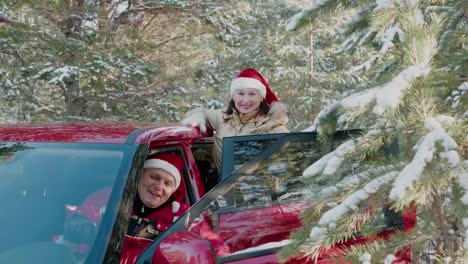 Fröhlicher-Mann-in-rotem-Weihnachtshut-wie-der-Weihnachtsmann-winkt-die-Hand-aus-dem-Auto-im-verschneiten-Wald.-Glückliches-Paar-im-Neujahr-Hut-im-Winterwald-auf-verschneiten-Bäumen-Landschaft.