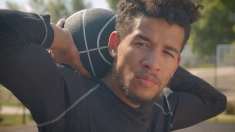 Closeup-portrait-of-young-attractive-african-american-male-basketball-player-looking-at-camera-holding-a-ball-behind-his-back-outdoors