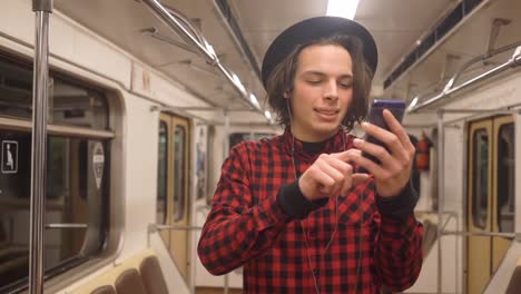 Young-handsome-stylish-man-wearing-black-hat-with-headphones-listening-to-music,-browsing-on-smartphone-in-public-transport,-millenial-hipster-boy-enjoying-mobile-technology-in-subway