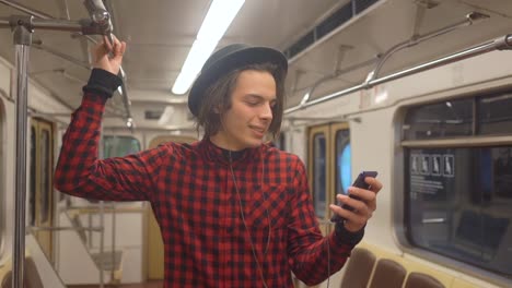 Young-handsome-stylish-man-wearing-black-hat-with-headphones-listening-to-music,-browsing-on-smartphone-in-public-transport,-millenial-hipster-boy-enjoying-mobile-technology-in-subway