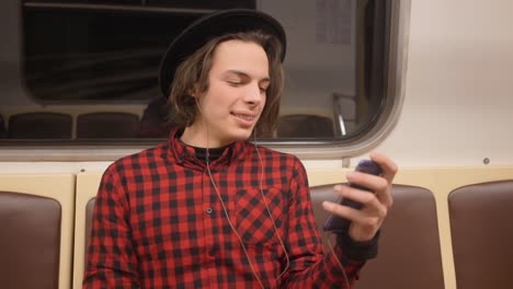 Young-handsome-stylish-man-wearing-black-hat-with-headphones-listening-to-music,-browsing-on-smartphone-in-public-transport,-millenial-hipster-boy-enjoying-mobile-technology-in-subway