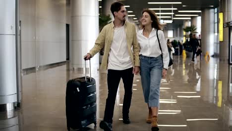 Vista-frontal-de-pareja-sonriente-caminando-juntos-en-el-aeropuerto-de-vacaciones-o-viaje.-Viajan-juntos.-Atractiva-mujer-caucásica-en-jeans-y-hombre-con-maleta-están-listos-para-viajar