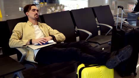 A-bearded-man-in-yellow-shirt-sleeping-holding-legs-on-the-yellow-suitcase,-holding-his-stuff-in-the-airport-lounge-while-sitting.-The-delay-of-the-flight